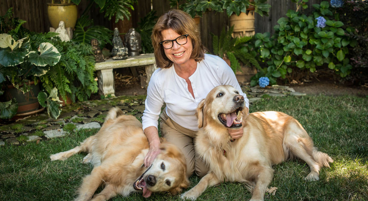 A woman smiling in the grass with her two golden retrievers