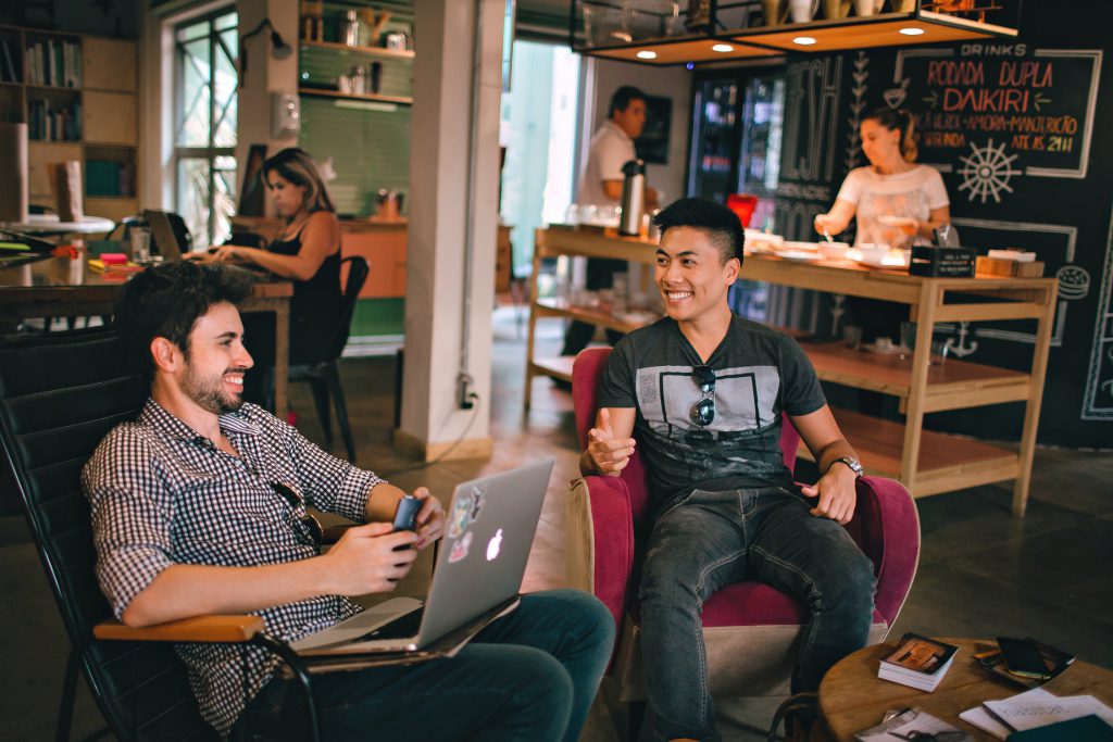 Two young men sitting smiling and interacting while sitting in chairs on a laptop in a coffeshop/lounge